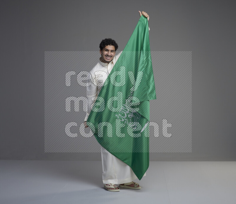 A saudi man standing wearing thob holding big saudi flag on gray background