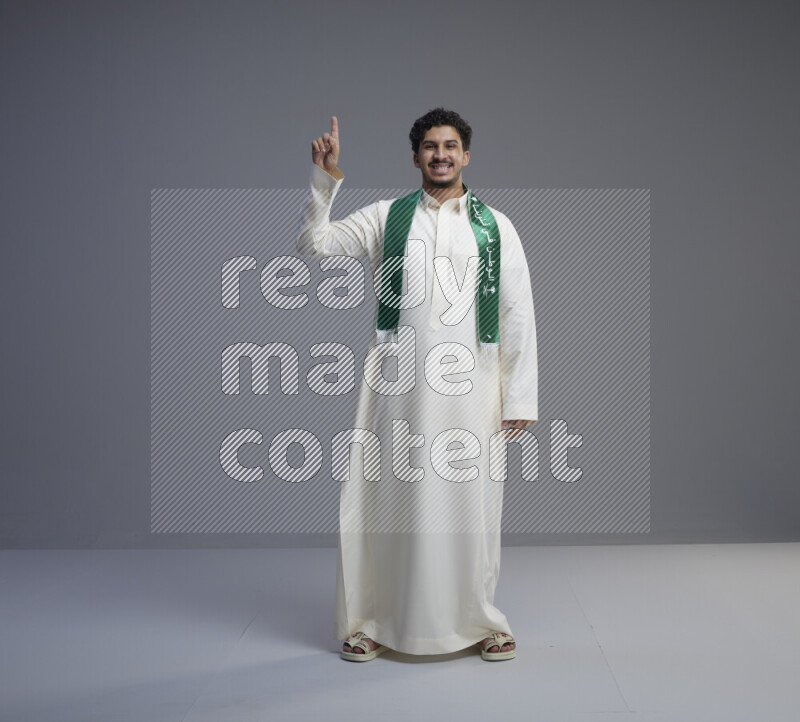 A Saudi man standing wearing thob and Saudi flag scarf on gray background