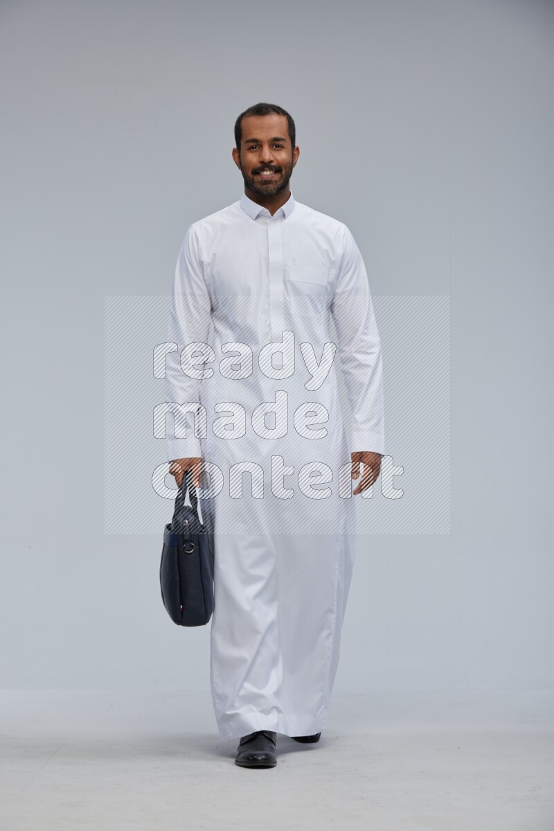 Saudi man wearing thob standing holding suitcase on gray background