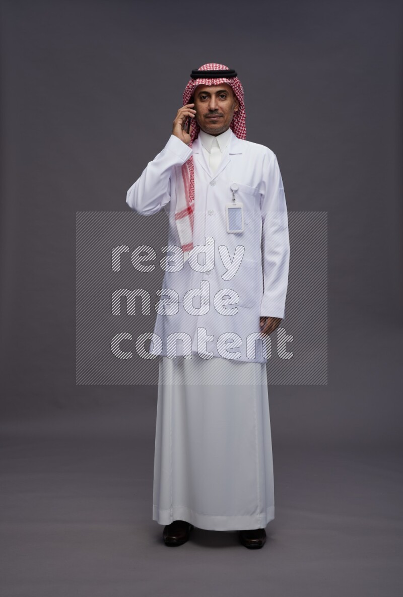 Saudi man wearing thob with lab coat and shomag with pocket employee badge standing talking on phone on gray background