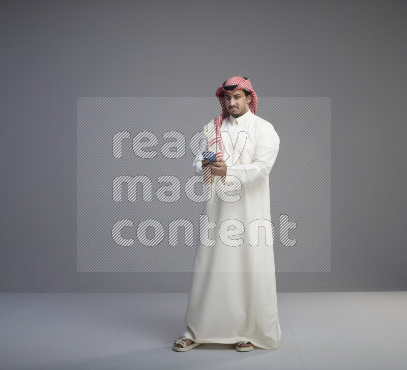 A  Saudi man standing wearing thob and red shomag texting on phone on gray background
