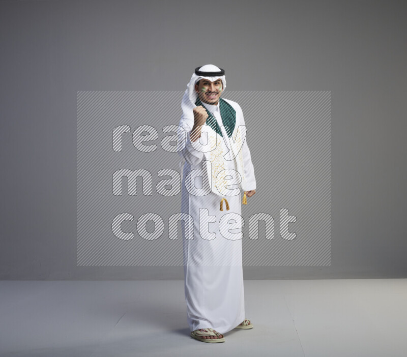 A Saudi man standing wearing thob and white shomag with face painting and Saudi flag scarf on gray background