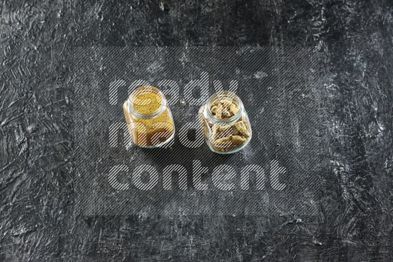 2 glass spice jars full of turmeric powder and dried whole fingers on textured black flooring
