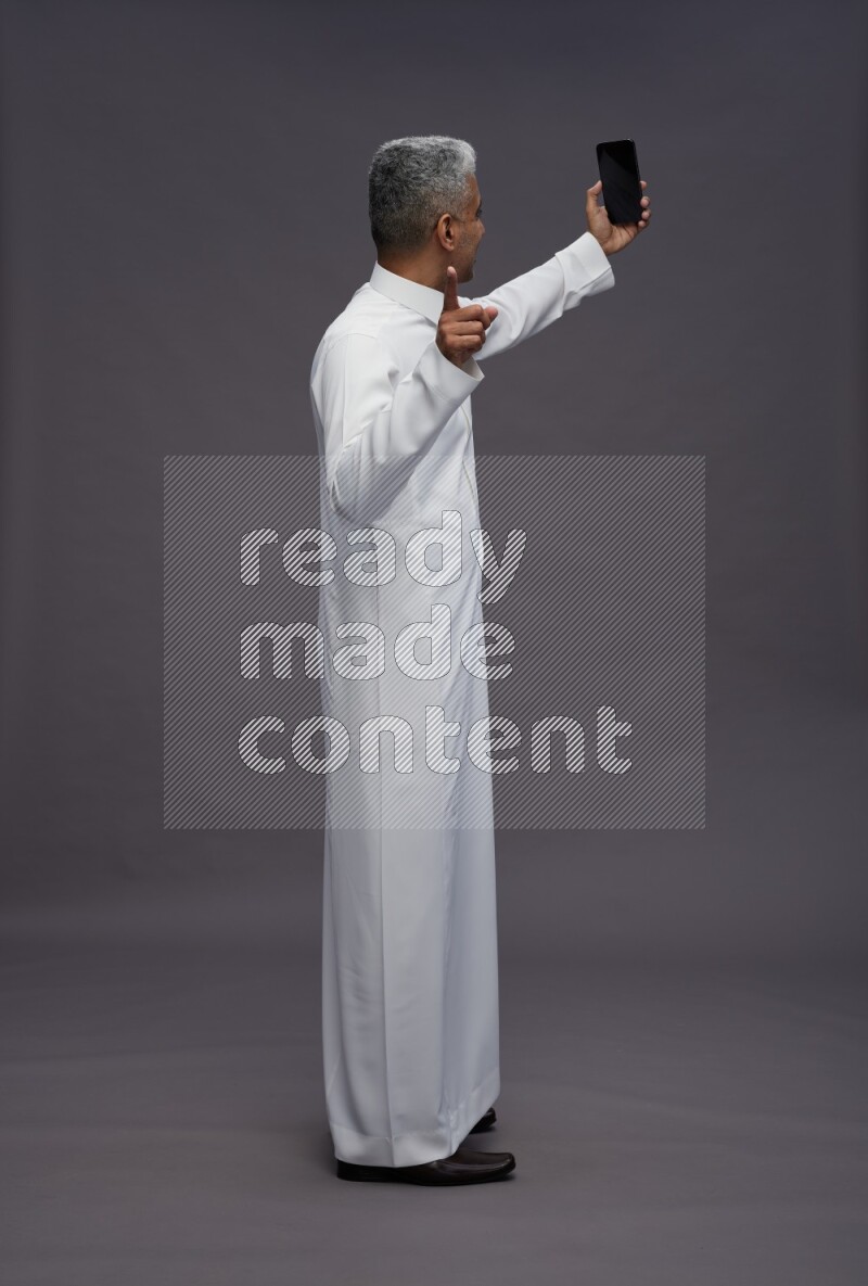 Saudi man wearing thob standing taking selfie on gray background