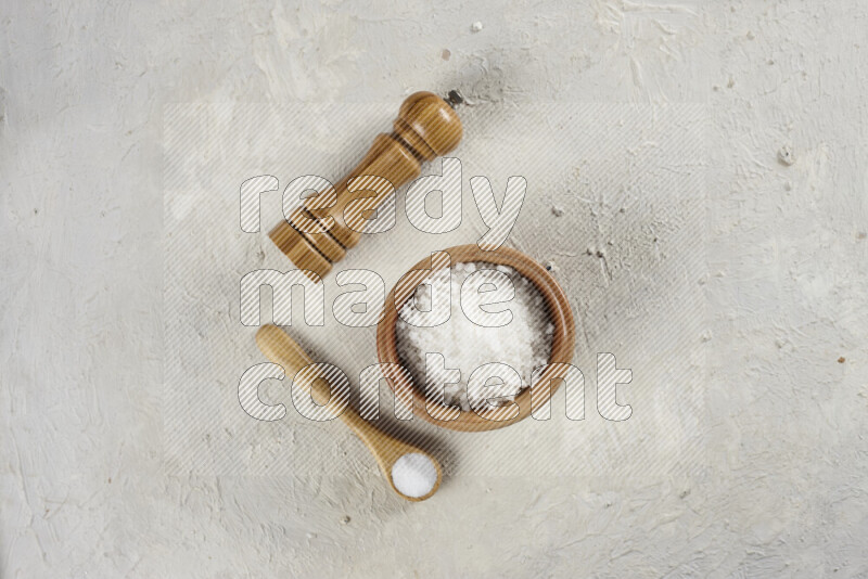 A wooden bowl and spoon filled with white sea salt and wooden grinder beside them on white background