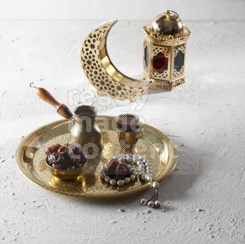 Dates in a metal bowl with coffee and prayer beads on a tray beside lanterns in a light setup
