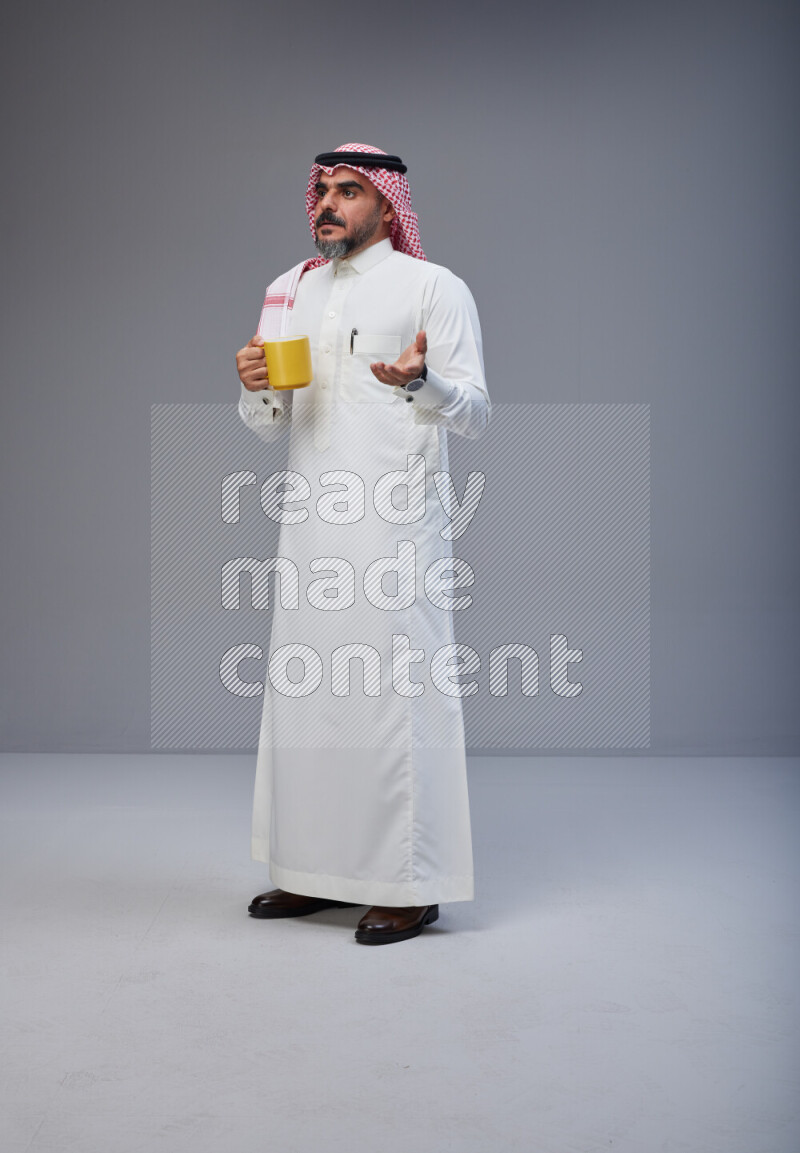 Saudi man Wearing Thob and red Shomag standing holding mug on Gray background