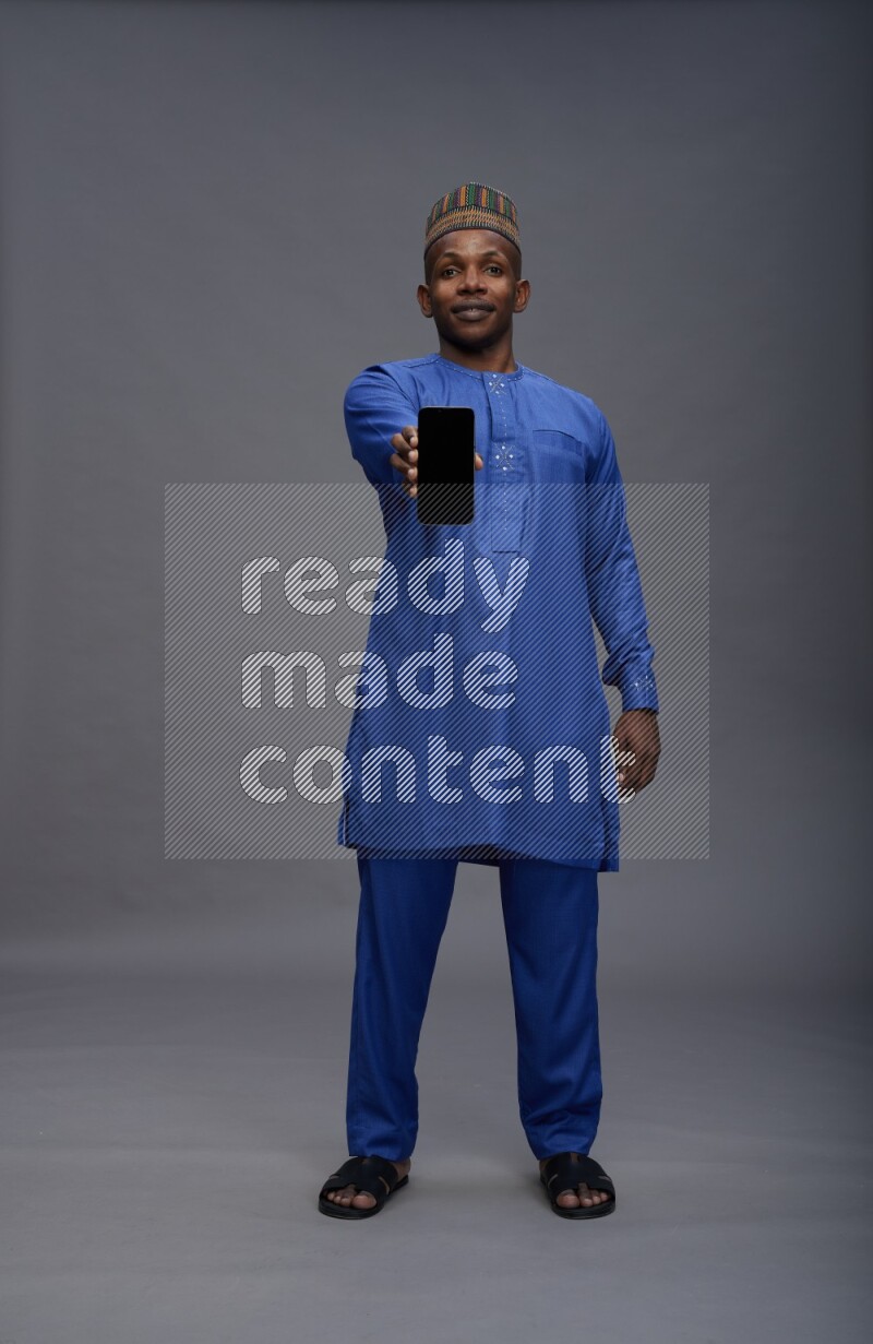 Man wearing Nigerian outfit standing showing phone to camera on gray background