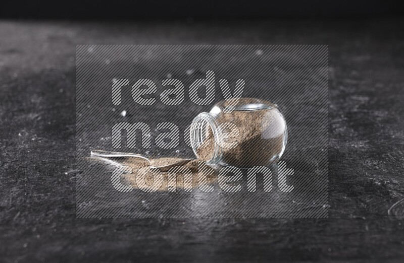 A flipped glass spice jar full of black pepper powder with a metal spoon full of powder on textured black flooring
