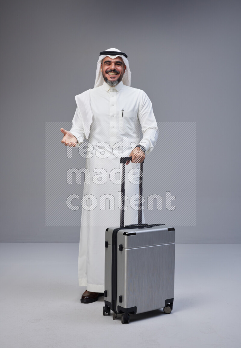 Saudi man wearing Thob and white Shomag standing holding Travel bag on Gray background