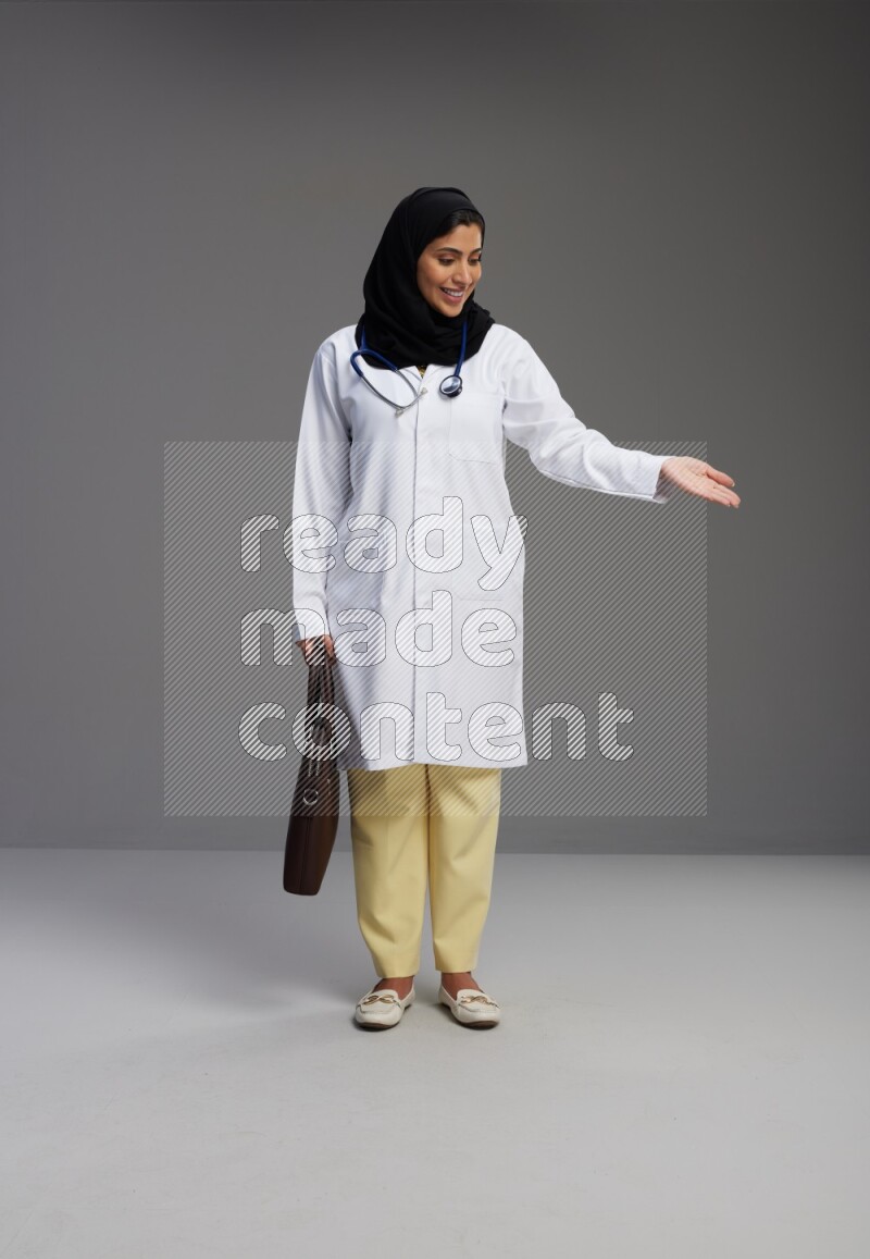 Saudi woman wearing lab coat with stethoscope standing holding bag on Gray background
