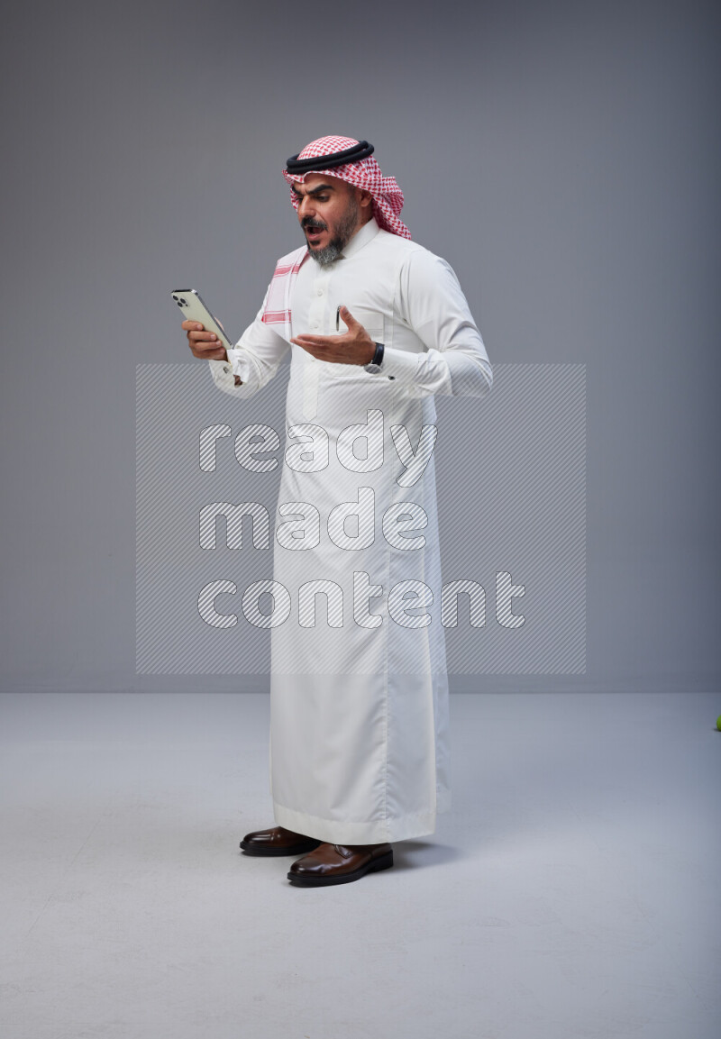 Saudi man Wearing Thob and red Shomag standing texting on phone on Gray background