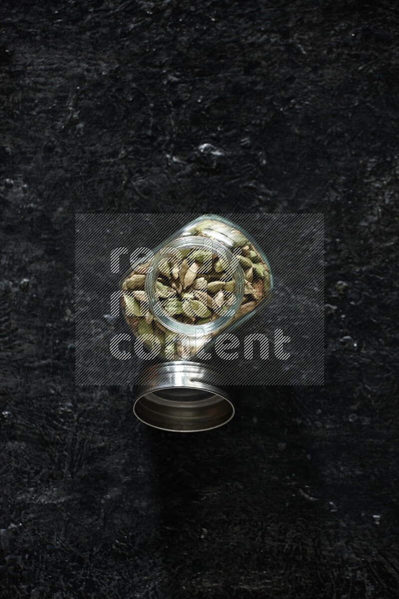 A glass spice jar full of cardamom seeds on textured black flooring