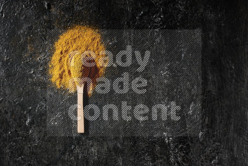 A wooden spoon full of turmeric powder on textured black background