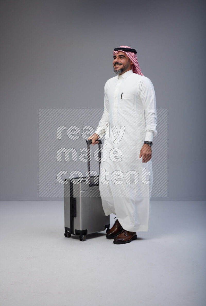 Saudi man wearing Thob and red Shomag standing holding Travel bag on Gray background