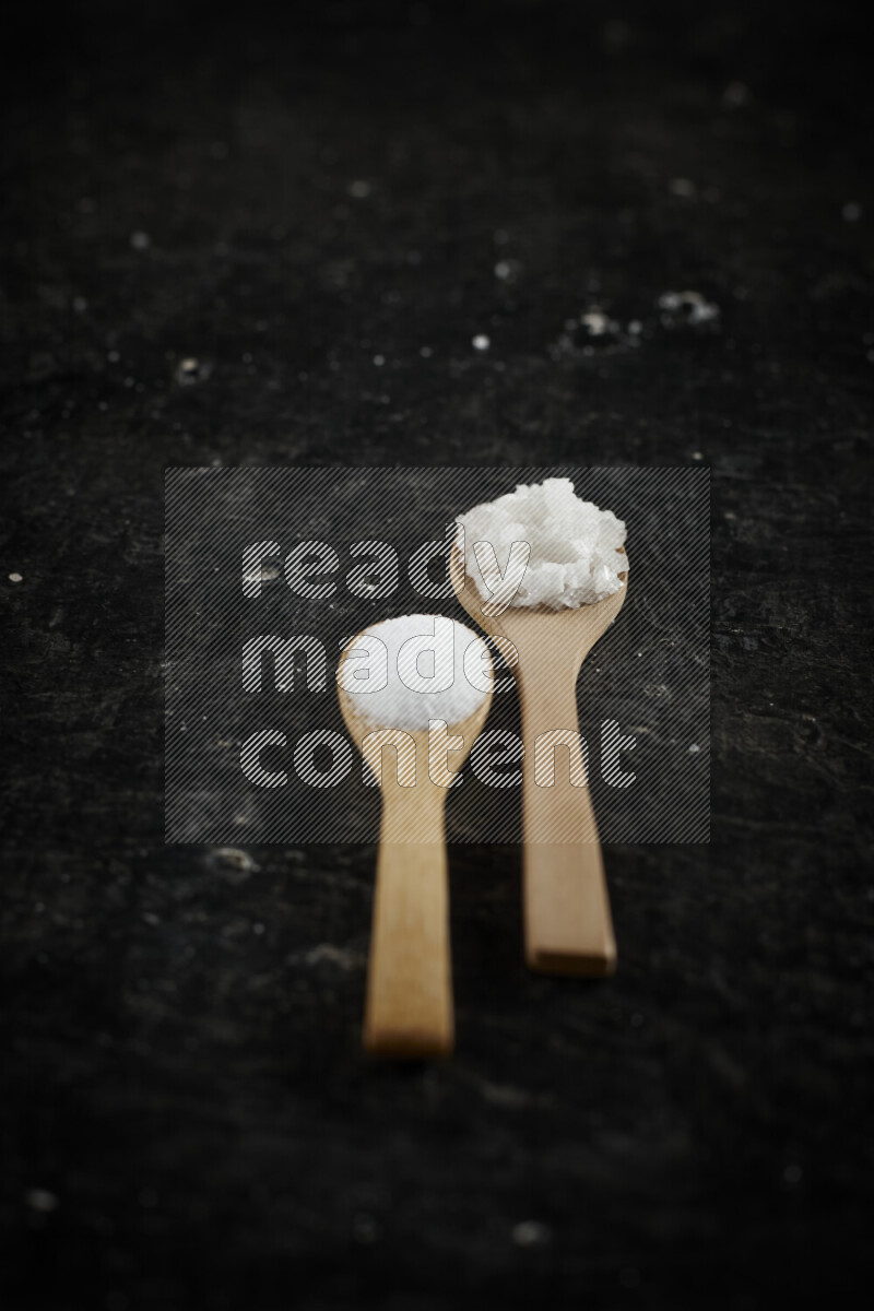 2 wooden spoons filled with fine and coarse salt on black background