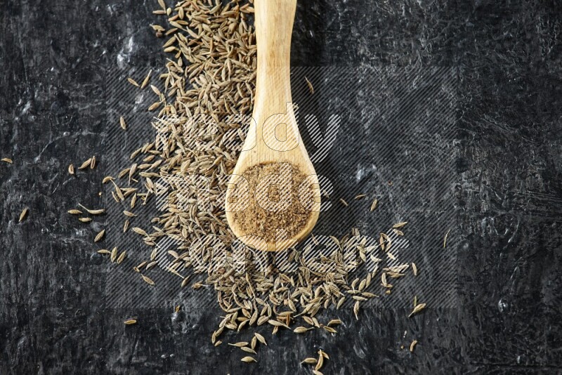 A wooden spoon full of cumin powder and cumin seeds spreaded on textured black flooring