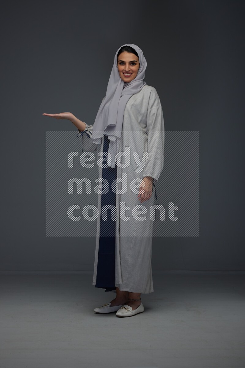 A Saudi woman wearing a light gray Abaya and head scarf standing and pointing in different directions eye level on a grey background