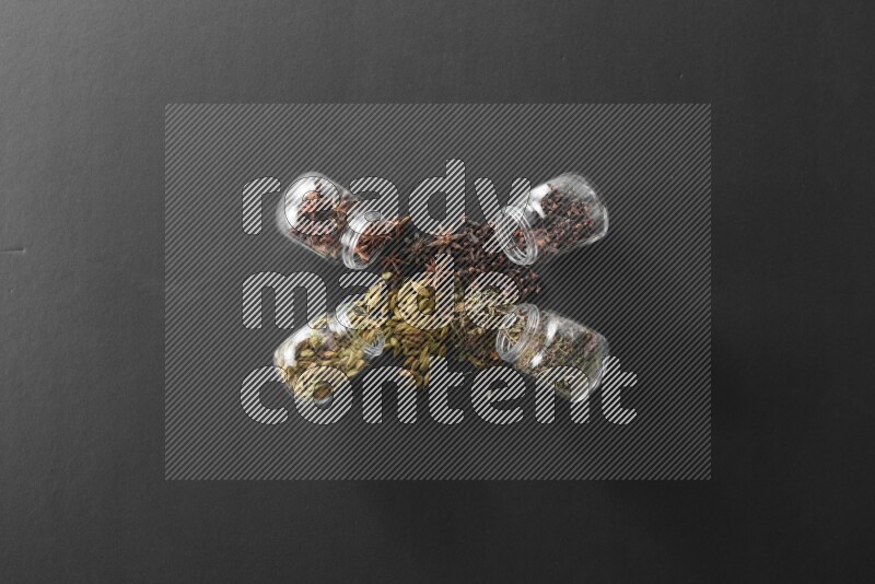 4 glass spice jars full of cloves, cardamom, basil and star anise on black flooring