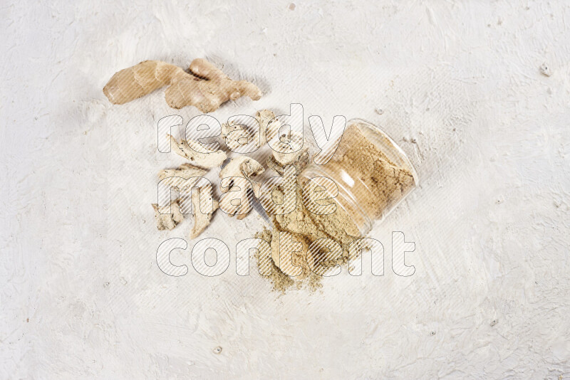 A glass jar full of ground ginger powder flipped with some spilling powder on white background