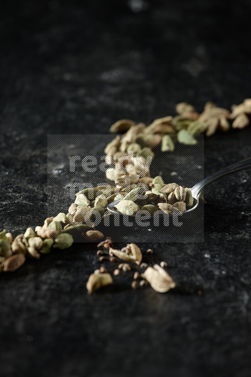 A Metal spoon full of cardamom seeds and some seeds beside it on a textured black flooring