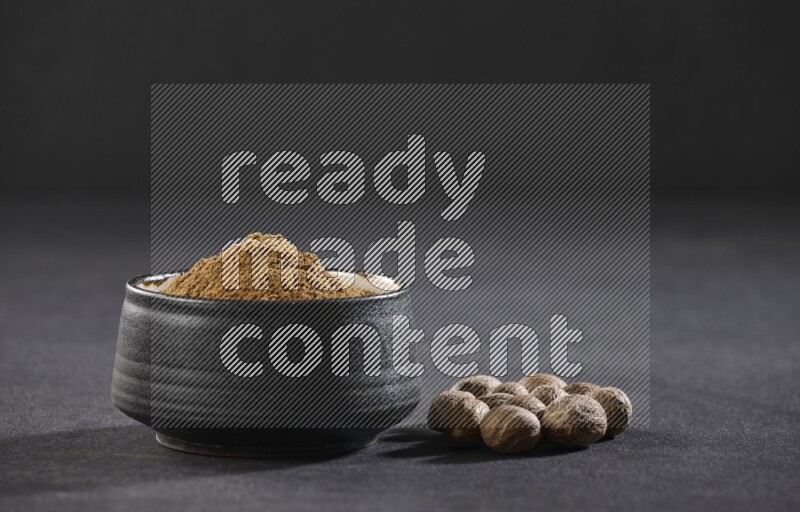 A black pottery bowl full of nutmeg powder with whole seeds beside it on a black flooring