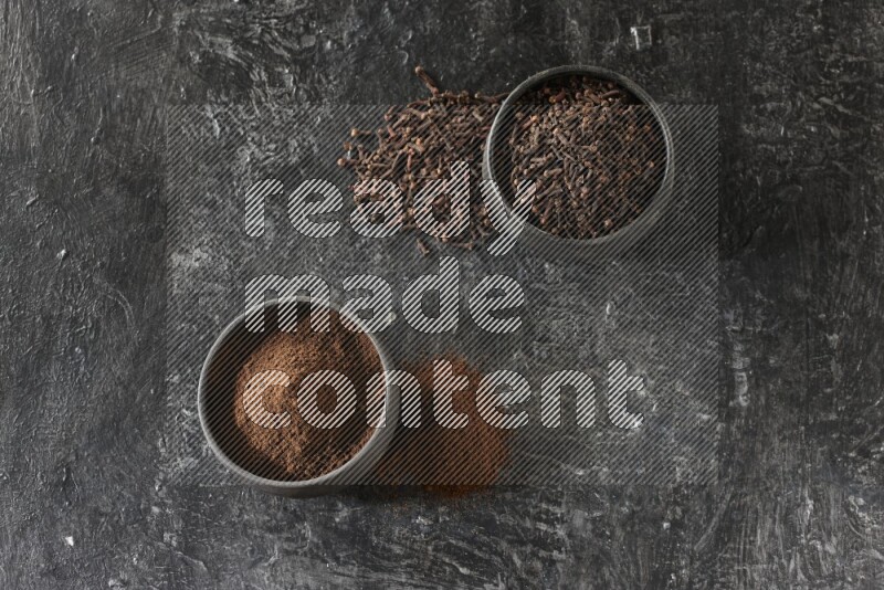 2 Black pottery bowls full of cloves and the other full of cloves powder on textured black flooring