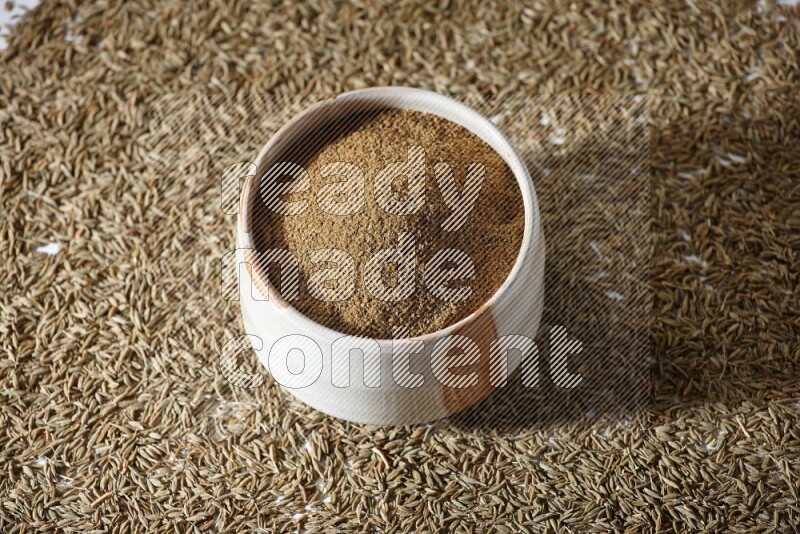 A beige bowl full of cumin powder with cumin seeds beneath it the bowl on a white flooring