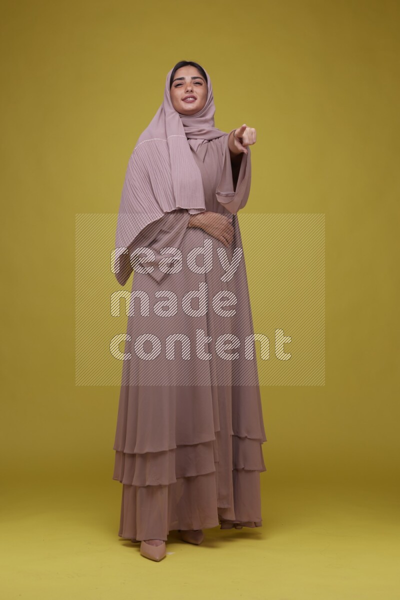 A woman Pointing on a Yellow Background wearing Brown Abaya with Hijab