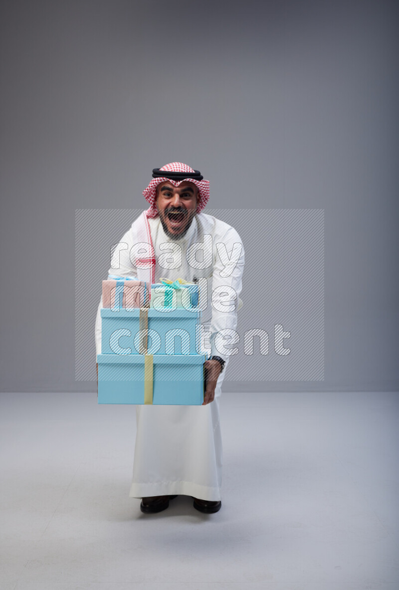 Saudi man Wearing Thob and red Shomag standing holding gift box on Gray background