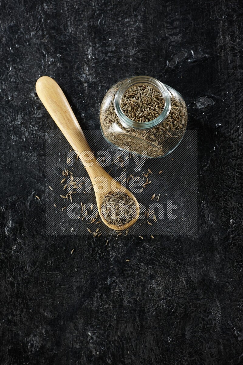 A glass spice jar and a wooden spoon full of cumin seeds on a textured black flooring
