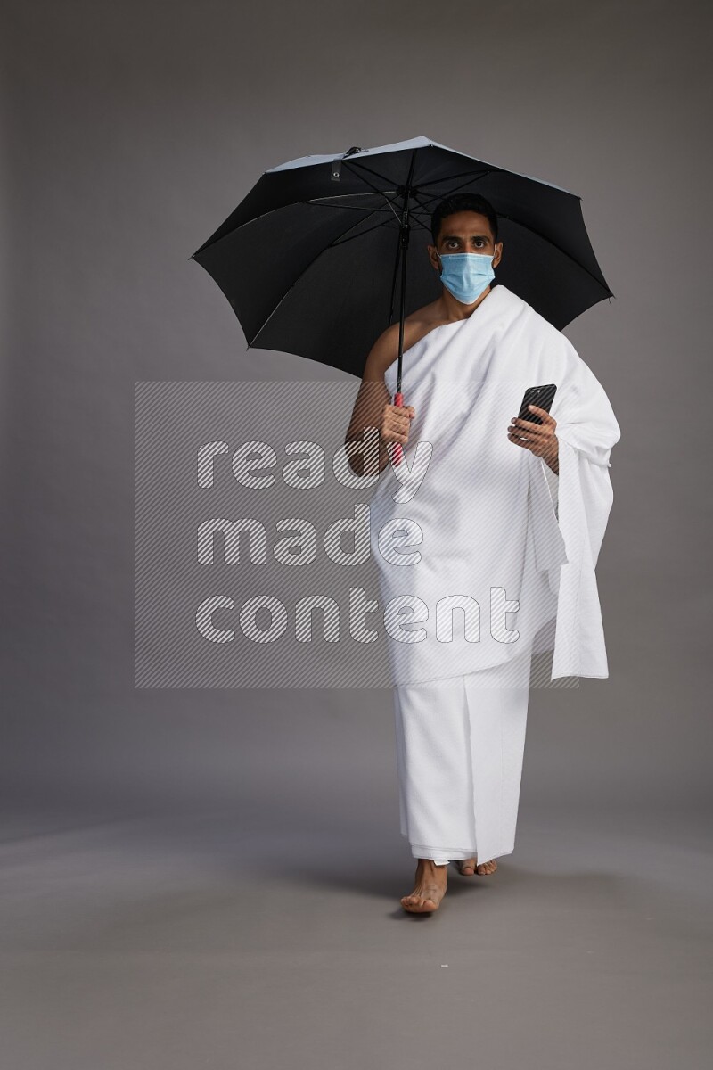 A man wearing Ehram and mask Standing holding umbrella on gray background on gray background