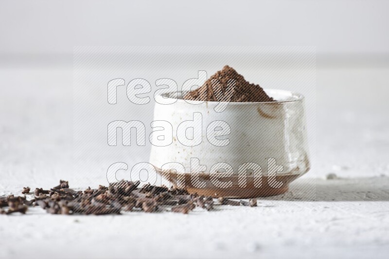 A beige ceramic bowl full of cloves powder and whole cloves on a white flooring
