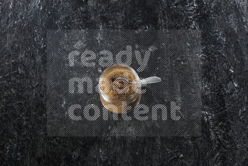 A glass jar and a metal spoon full of cumin powder on a textured black flooring