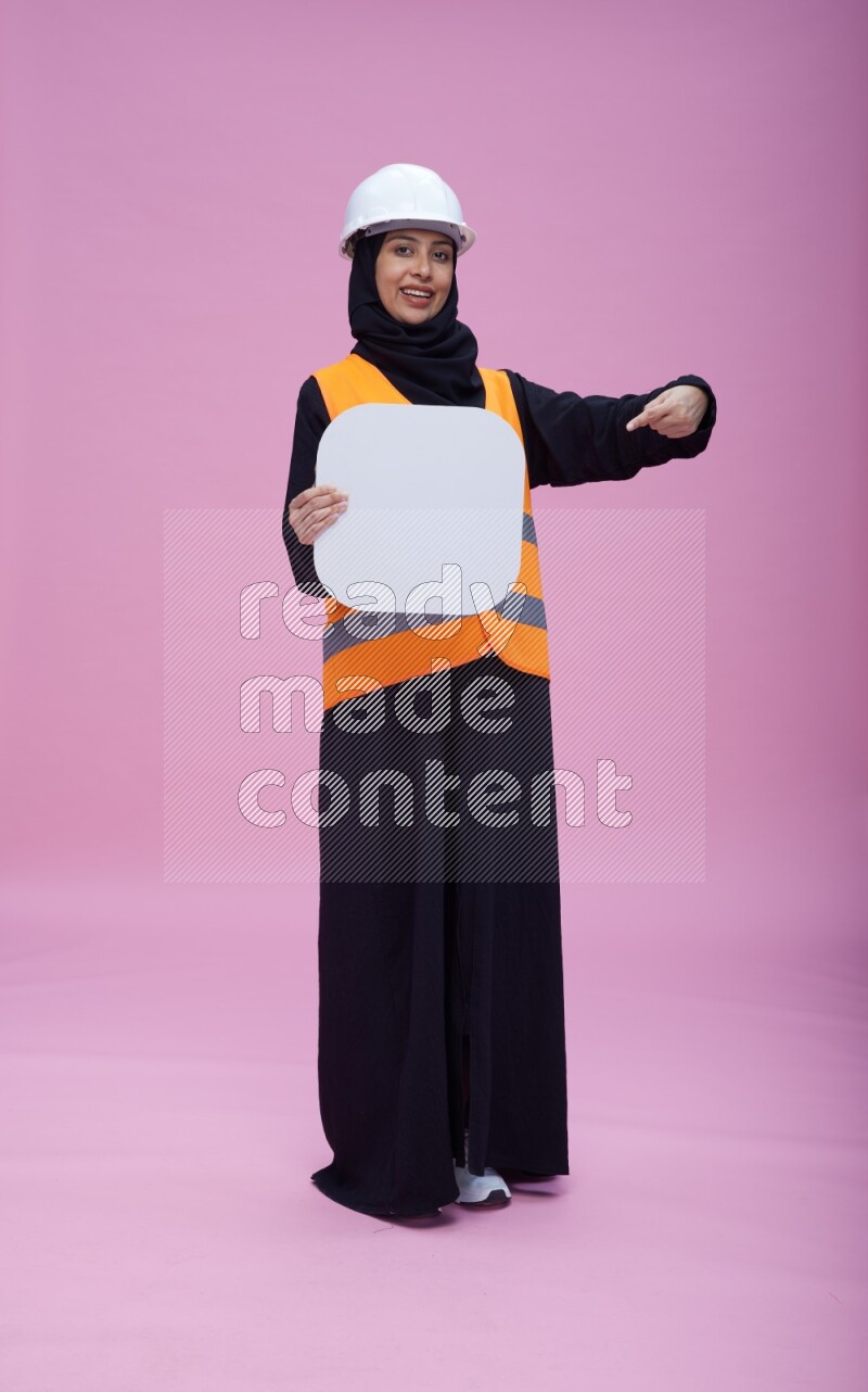 Saudi woman wearing Abaya with engineer vest and helmet standing holding social media sign on pink background