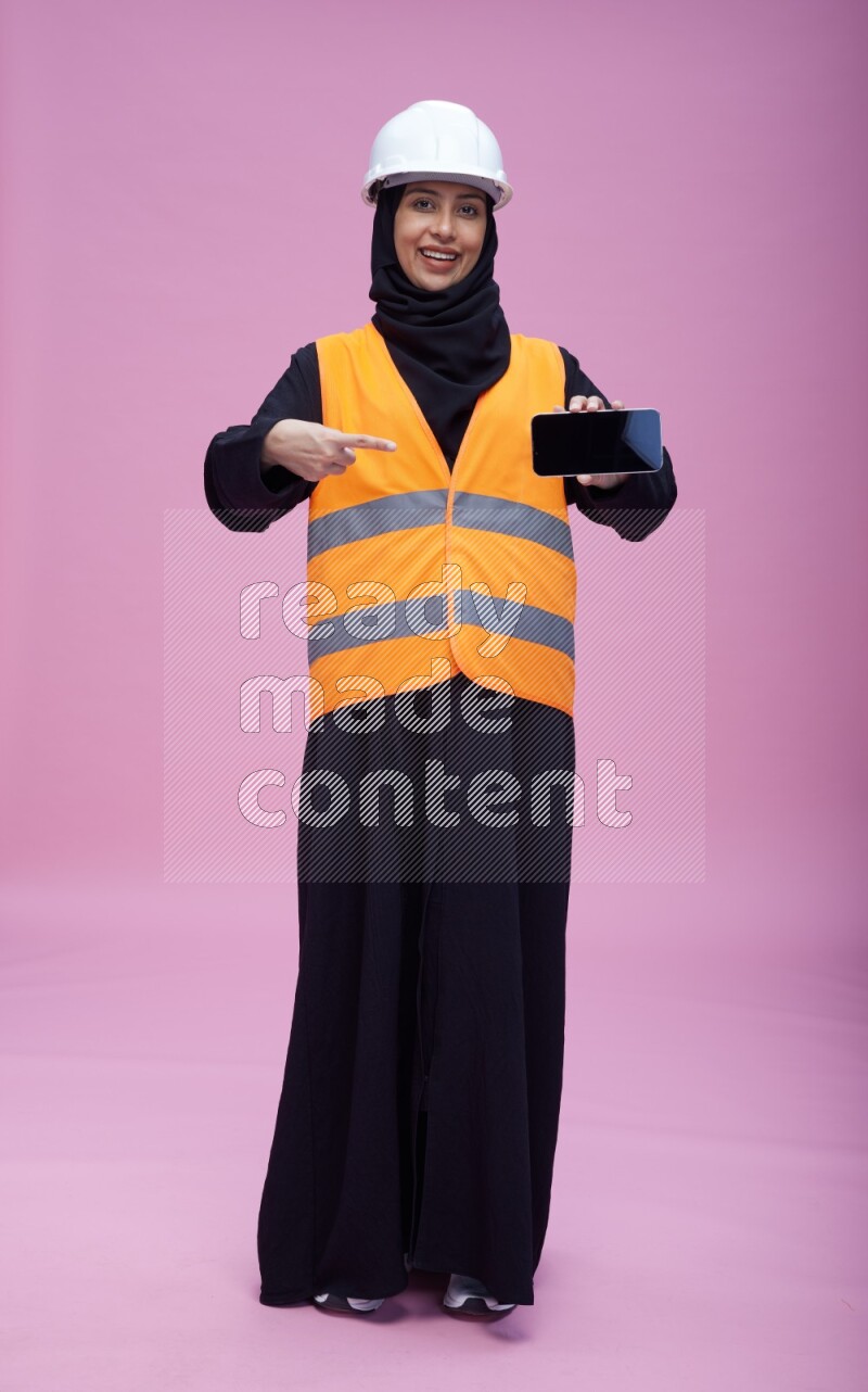Saudi woman wearing Abaya with engineer vest and helmet standing showing phone to camera on pink background