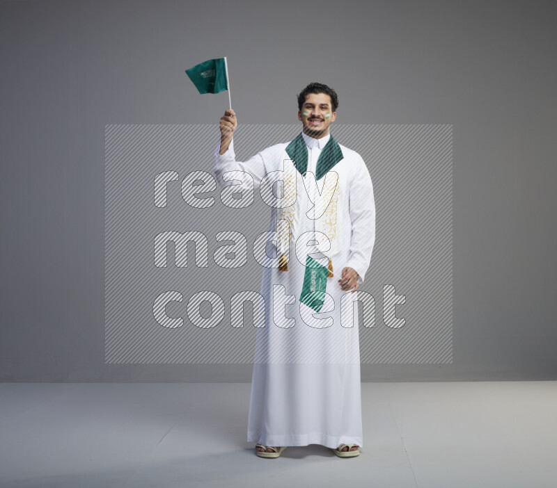 A Saudi man standing wearing thob and saudi flag scarf with face painting holding small Saudi flag on gray background