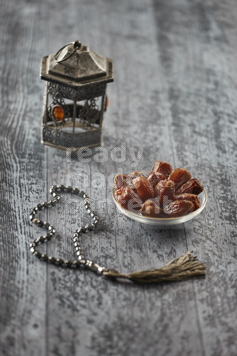 A silver lantern with different drinks, dates, nuts, prayer beads and quran on grey wooden background