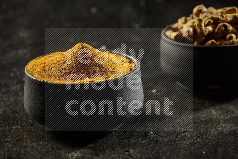 2 pottery black bowls, one full of turmeric powder and the other full of dried turmeric whole fingers on a textured black flooring