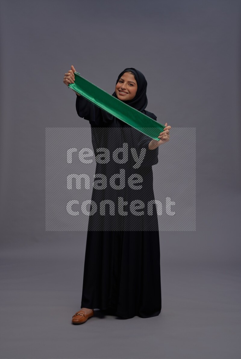 Saudi woman wearing Abaya standing holding Saudi flag on gray background