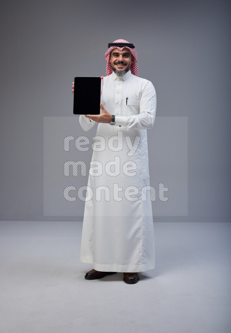 Saudi man Wearing Thob and red Shomag standing showing tablet to camera on Gray background