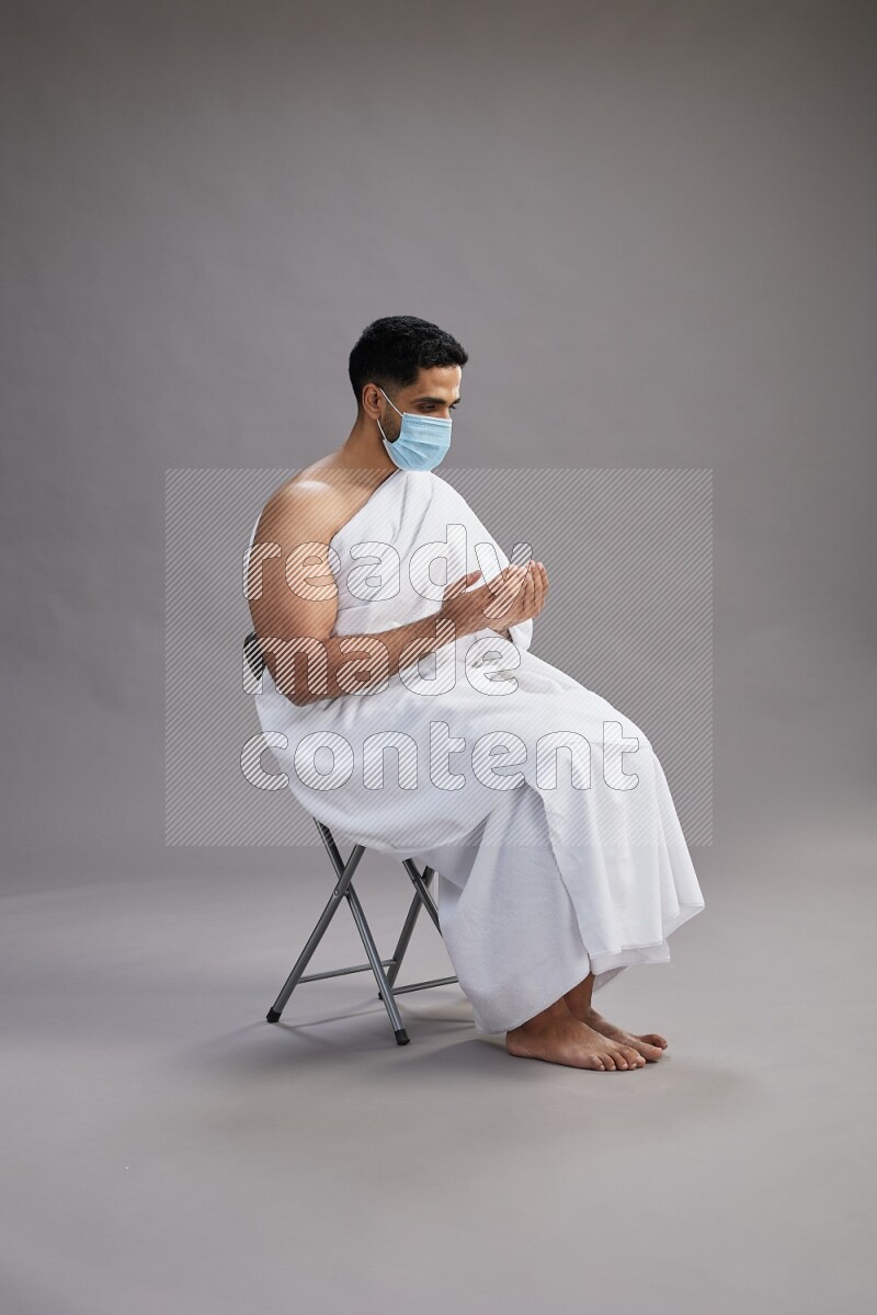 A man wearing Ehram with face mask sitting on chair dua'a on gray background