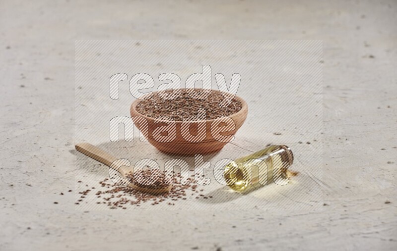 A wooden bowl and spoon full of flaxseeds with a bottle of flaxseeds oil on a textured white flooring