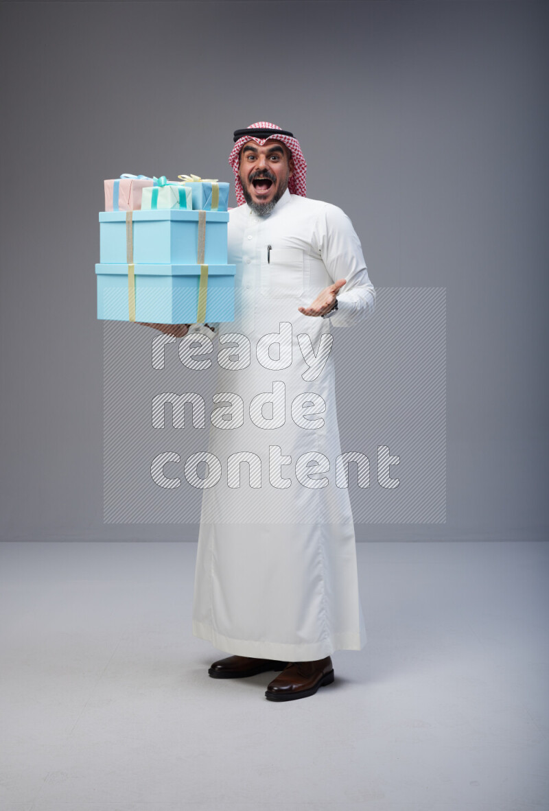 Saudi man Wearing Thob and red Shomag standing holding gift box on Gray background