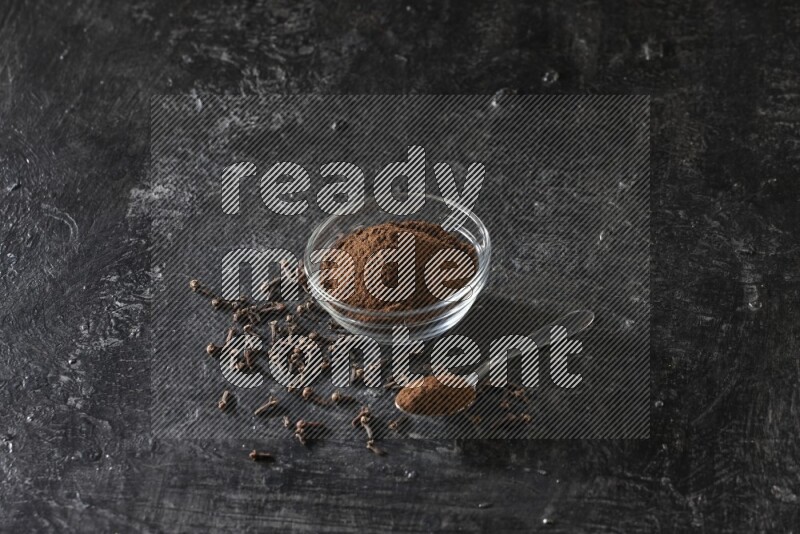 A glass bowl and a metal spoon full of cloves powder with gloves grains beside them on a textured black flooring