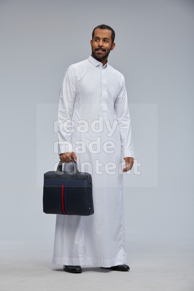 Saudi man wearing thob standing holding suitcase on gray background