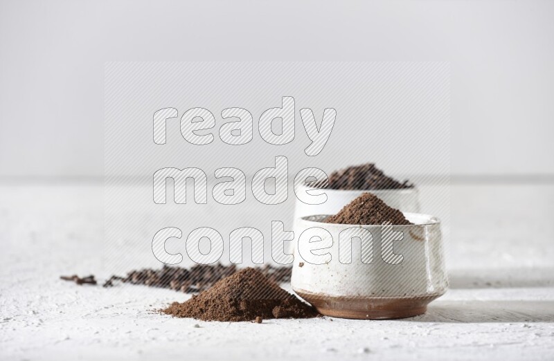 2 Beige ceramic bowls, one full of cloves and the other full of powder on textured white flooring