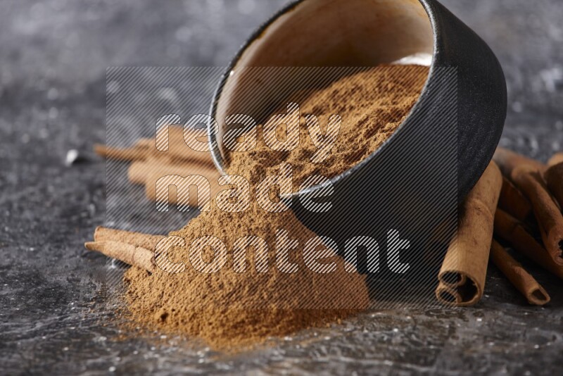 Black pottery bowl over filled with cinnamon powder and cinnamon sticks around the bowl on a textured black background