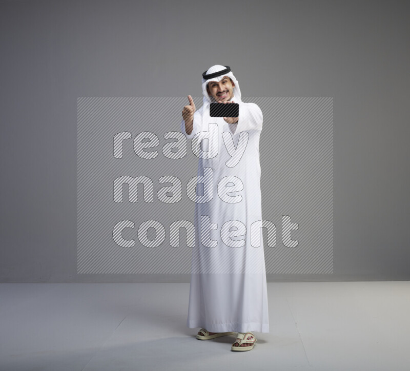 A Saudi man standing wearing thob and white shomag showing phone to camera on gray background