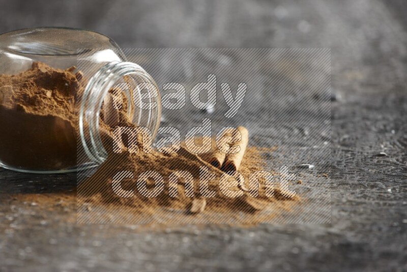 Flipped glass herbs jar full of cinnamon powder with cracked cinnamon sticks on a textured black background
